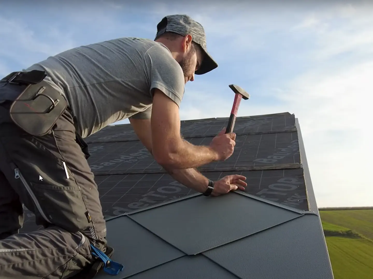 Spengler in Aktion: Mit einem Hammer verlegt der Eigentümer der Firma „Der Blechmann“ präzise die PREFA Dachraute 44 × 44. Die Präzision bei der Installation ist bei den bereits verlegten Rauten am unteren Bildrand zu erkennen. Der Mann trägt eine Mütze und kniet mit dem Rücken zur Kamera auf dem Dach. Im Hintergrund ist der klare Himmel und eine grüne Landschaft zu erkennen. Der harmonische Hintergrund unterstreicht die Symbolik des Bildes: Nachhaltiges und langlebiges Handwerk mit hochwertigen PREFA Aluminiumprodukten.