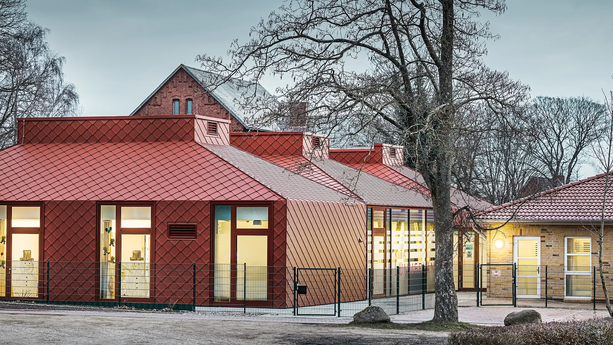 Modernes Kindergartengebäude mit auffälliger Fassade und Dacheindeckung aus rautenförmigen Aluminiumplatten in Oxydrot. Die Architektur kombiniert traditionelle und moderne Elemente mit großen Fenstern und klaren Linien. Das Gebäude ist von Bäumen umgeben, im Vordergrund verläuft ein Zaun, und die Beleuchtung im Inneren schafft eine warme Atmosphäre in der Dämmerung.