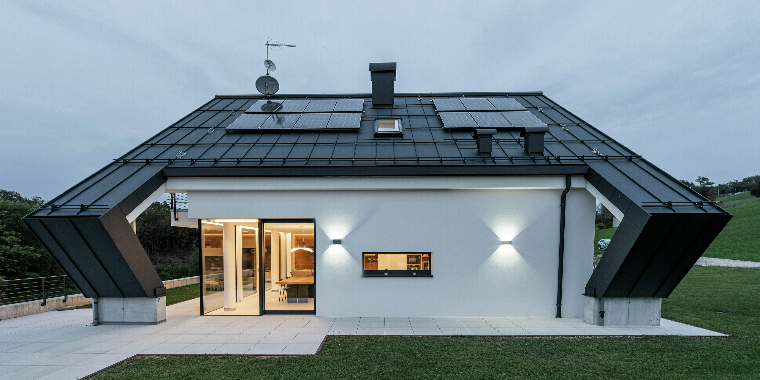Modernes Einfamilienhaus in Borgo Valbelluna, Italien, mit markantem Satteldach aus PREFALZ in P.10 Anthrazit. Die langlebige Aluminium-Dacheindeckung bietet optimalen Schutz vor Witterungseinflüssen und überzeugt durch flexible Verlegemöglichkeiten. Integrierte Photovoltaikmodule nutzen die Dachfläche effizient zur nachhaltigen Energiegewinnung. Die Kombination aus anthrazitfarbenem Aluminiumdach und weißer Fassade sowie die markante Auskragung des Satteldaches unterstreichen das elegante und zeitlose Design des Gebäudes.