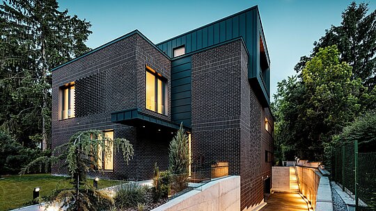 Die Aufnahme zeigt die moderne, klar strukturierte Außenansicht der Budapester Mehrgenerationen‑Villa in der Abendstimmung. Dunkelrotbraune Backsteinfassaden treffen auf präzise ausgeführte PREFALZ Aluminiumverkleidungen in P.10 Anthrazit, die durch ihre vertikalen Linien einen starken architektonischen Akzent setzen. Warm erleuchtete Fensteröffnungen verleihen dem Gebäude Tiefe und Atmosphäre. Die sanft beleuchtete Zufahrt zur Tiefgarage führt elegant am Baukörper entlang und betont die hochwertige, langlebige und wetterbeständige Materialkombination aus Klinker und PREFA Aluminium.