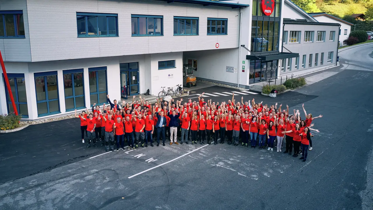 PREFA MitarbeiterInnen mit rotem PREFA T-Shirt stehen in einem Halbkreis vor dem Bürogebäude in Marktl.