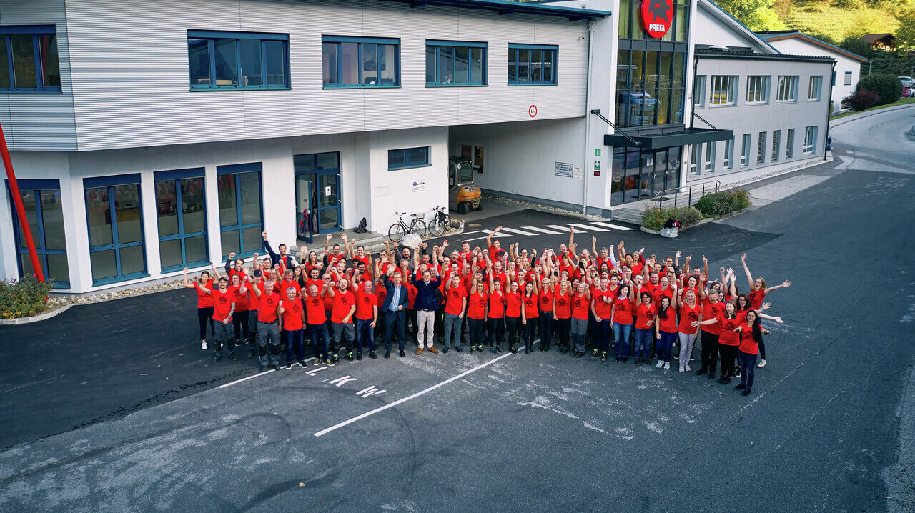 PREFA MitarbeiterInnen mit rotem PREFA T-Shirt stehen in einem Halbkreis vor dem Bürogebäude in Marktl.