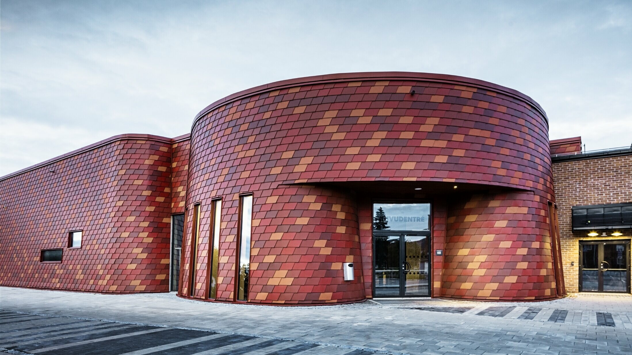 Eishalle mit Flachdach und geschwungener Fassade, die Fassade ist mit Wandschindeln aus Aluminium von PREFA in verschiedenen Rottönen verkleidet - Oxydrot, Ziegelrot, Rotbraun, Braunrot