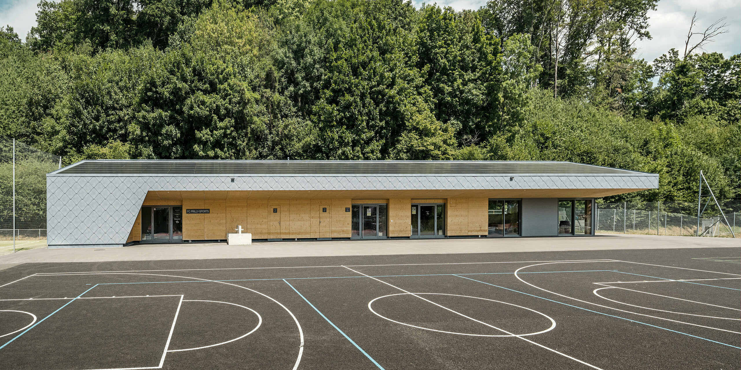 Frontansicht der Sportanlage Fleur de Lys in Prilly, Schweiz, mit einer hochwertigen Verkleidung aus PREFA Dach- und Wandrauten 44x44 in P.10 Steingrau. Das Bild zeigt das moderne, eingeschossige Gebäude mit seiner klaren Linienführung und der harmonischen Integration in die umgebende Natur. Die Sportanlage steht vor einem asphaltiertem Basketballplatz, der auch für andere Sportarten wie Kleinfeldfußball genutzt wird. Die Verwendung von PREFA Aluminiumprodukten untermauert die Beständigkeit und Ästhetik des Bauwerks, während es gleichzeitig moderne Architektur und nachhaltige Bauweisen repräsentiert. Die überdachten Bereiche der Sportanlage wurden mit Holzelementen verkleidet, was die Synergie zwischen Holz und Aluminium unterstreicht.