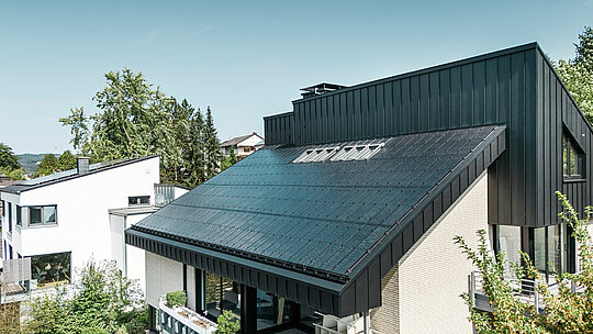 Moderne Architektur eines Einfamilienhauses in Meschede, Deutschland, mit einem PREFALZ Aluminiumdach in P.10 Schwarz und integrierten Glas/Glas PV-Modulen. Das versetzte Pultdach verbindet zeitloses Design mit nachhaltiger Photovoltaik-Technologie. Die innovativen PREFALZ Solarmodule liefern eine Leistung von 18 kWp. Die rahmenlosen Module und die elegante Aluminiumverkleidung von Fassade und Dach bilden eine ästhetische und funktionale Einheit, die sich perfekt in das grüne Wohnumfeld einfügt.