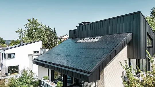 Moderne Architektur eines Einfamilienhauses in Meschede, Deutschland, mit einem PREFALZ-Aluminiumdach in P.10 Schwarz und integrierten Glas/Glas-Photovoltaikmodulen. Das versetzte Dach vereint zeitloses Design mit nachhaltiger Photovoltaik-Technologie. Die innovativen PREFALZ-Solarmodule liefern eine Leistung von 18 kWp. Die rahmenlosen Module und die elegante Aluminium-Dach- und Fassadenverkleidung bilden ein ästhetisches und funktionales Ganzes, das sich harmonisch in die grüne Wohnumgebung einfügt.