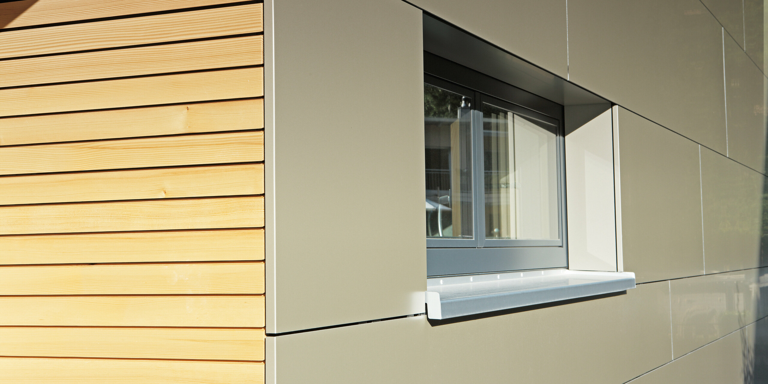 Detailaufnahme der Fassade eines Einfamilienhauses in Hohenems, die eine harmonische Kombination aus PREFA Aluminium Verbundplatten in Bronzemetallic und einer natürlichen Lärchenholzverkleidung zeigt. Das Bild fängt die feine Textur des Holzes und die glatte Oberfläche der Verbundplatten ein, die zusammen für eine attraktive Optik sorgen. Die hochwertigen Materialien reflektieren die Sonne und betonen die klare, moderne Ästhetik des Hauses.