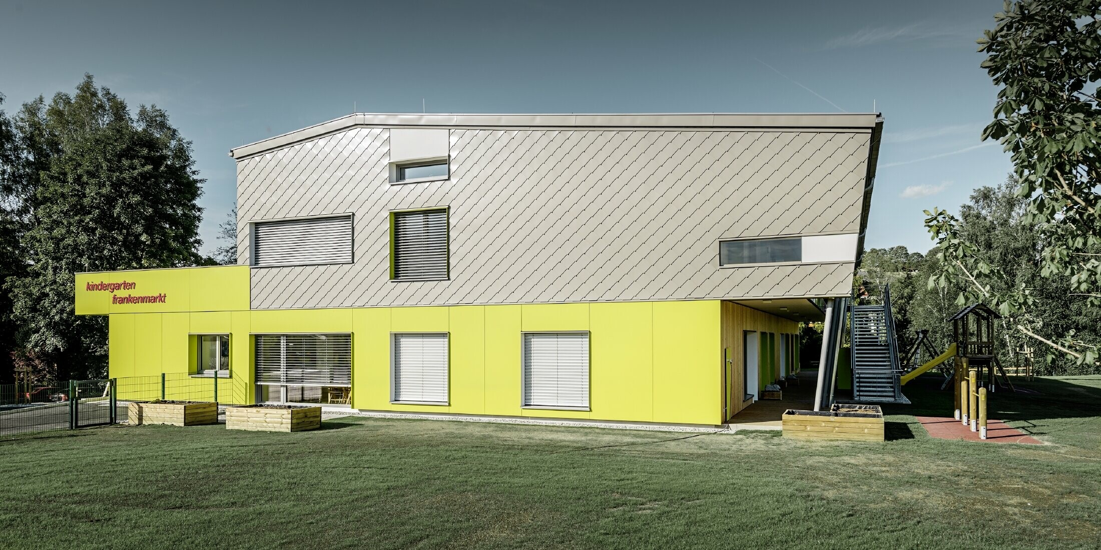 Der Kindergarten wurde teilweise mit der PREFA Wandraute 29 in Bronze verkleidet, dieser Gebäudeteil hat auch keinen Dachvorsprung. Der Rest der Fassade wurde mit Fassadenplatten in Gelb verkleidet.