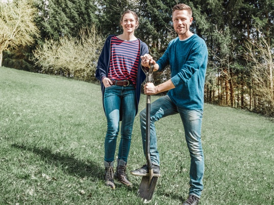 Junges Pärchen steht auf ihrem noch unbebauten Baugrund. Der Mann hält einen Spaten in der Hand. In dieser Bauphase holen sich die Bauherren Angebote für den Hausbau.