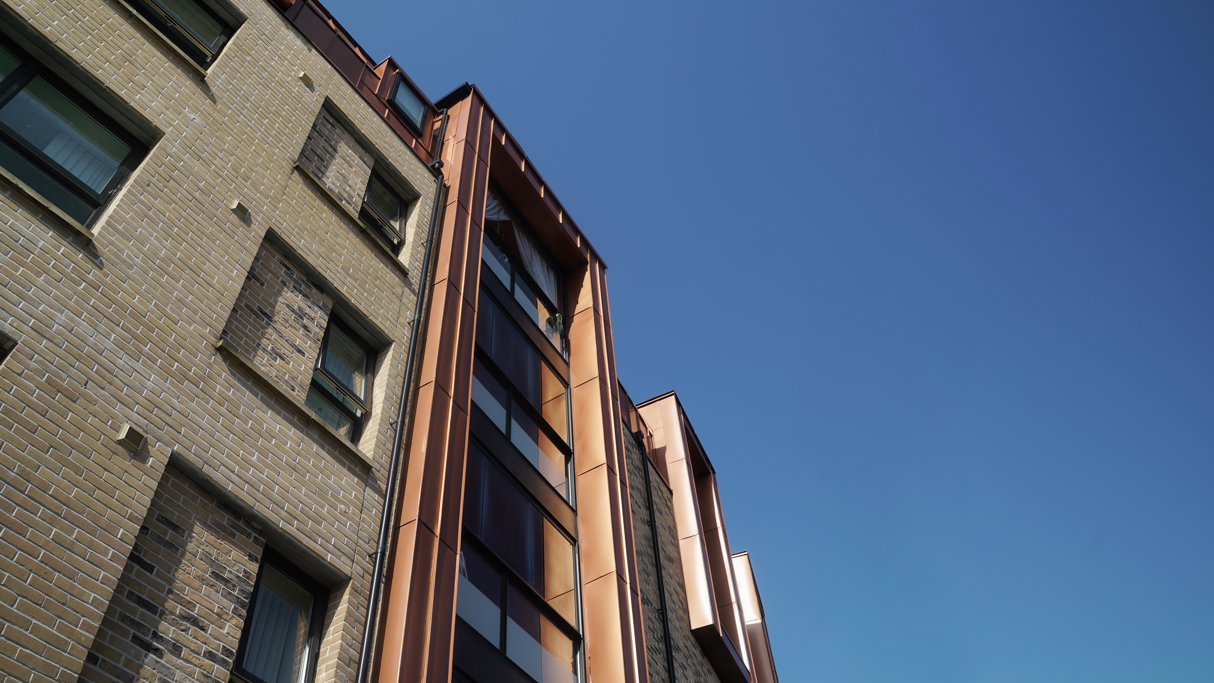 Perspektivische Ansicht der Fassade einer modernen Wohnhausanlage in der Nethan Street in Glasgow, verkleidet mit FALZONAL in Neukupfer. Die metallischen Fassadenelemente fangen das Licht ein und reflektierten es, was die Gebäudeform betont und einen eindrucksvollen visuellen Kontrast zur traditionellen Backsteinarchitektur bietet. Das PREFA Aluminiumprodukt sorgt nicht nur für eine ansprechende Ästhetik, sondern verspricht auch Langlebigkeit und geringen Wartungsaufwand.