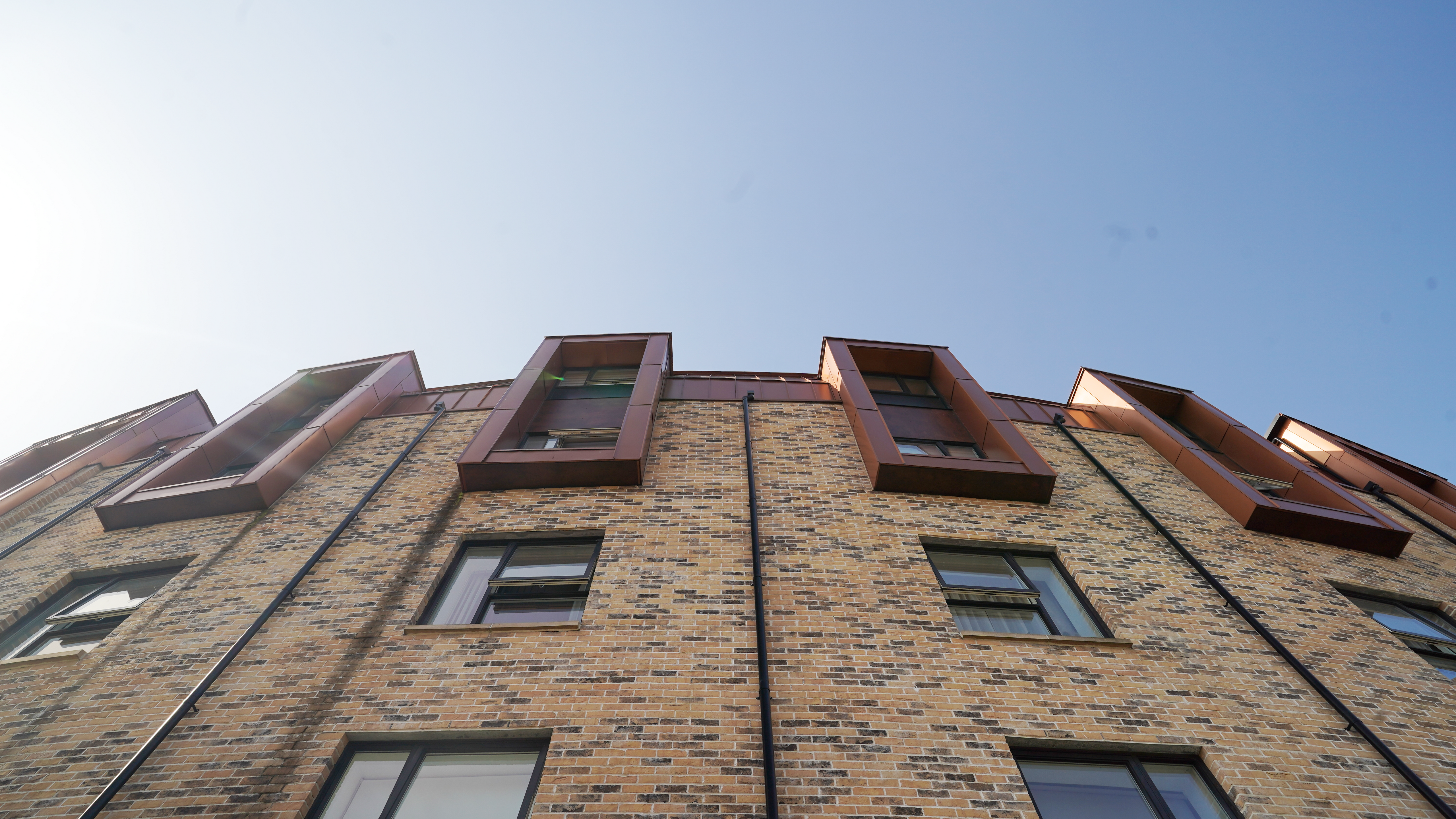 Aufwärtsblick auf die Fassade einer Wohnhausanlage in der Nethan Street in Glasgow, die sich durch die Verwendung von FALZONAL in Neukupfer auszeichnet, einem langlebigen und robusten PREFA Aluminiumprodukt. Die vertikale Ausrichtung der Fenster, die mit dem kupferfarbenen Aluminium umrahmt wurden, unterbricht die horizontale Gliederung der Ziegelmauer und verleiht dem Gebäude eine dynamische und moderne Ausstrahlung. Die Qualität und Farbgebung des FALZONAL fügt sich nahtlos in die städtische Architektur ein und bietet gleichzeitig eine beständige und ästhetische Lösung für moderne Wohnkomplexe. Zwischen den Fenstern sind schwarze Ablaufrohre zur Dachentwässerung platziert worden, die in dieser Perspektive die Rundung des Bauwerks verdeutlichen.